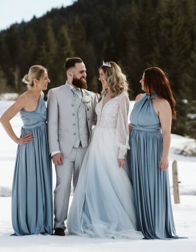 Brautpaar mit Brautjungfern und luxuriösen Blumengestecken im Schnee bei ihrer Winterhochzeit in den Bergen