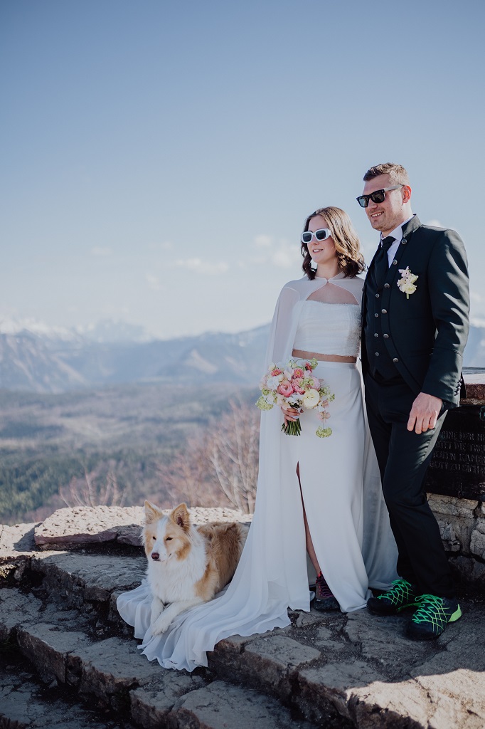 Bridal couple with their dog at the summit cross during the photo shoot at their mountain wedding in Austria