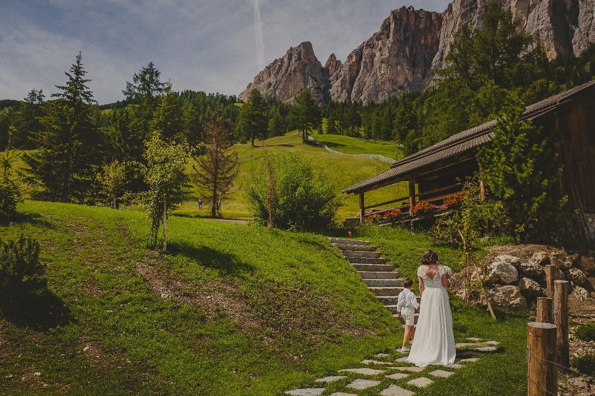 Braut mit Kind auf dem Gartenweg unterhalb des Kolfuschgerhofs – natürliche und emotionale Szene bei einer Sommerhochzeit in den Dolomiten.